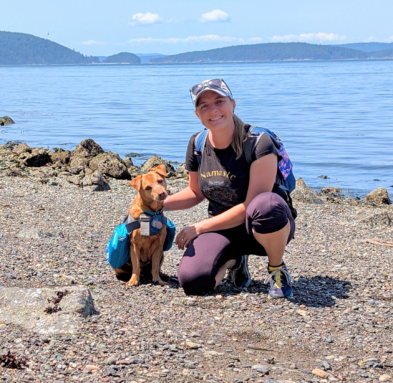 Rachel kneeling with Maui on a rocky beach at Cypress Island with the San Juan Islands in the background