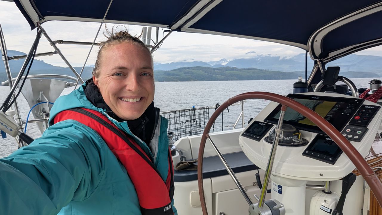 Rachel smiling at the helm of SV Yahara in Johnstone Strait with snow-capped mountains behind