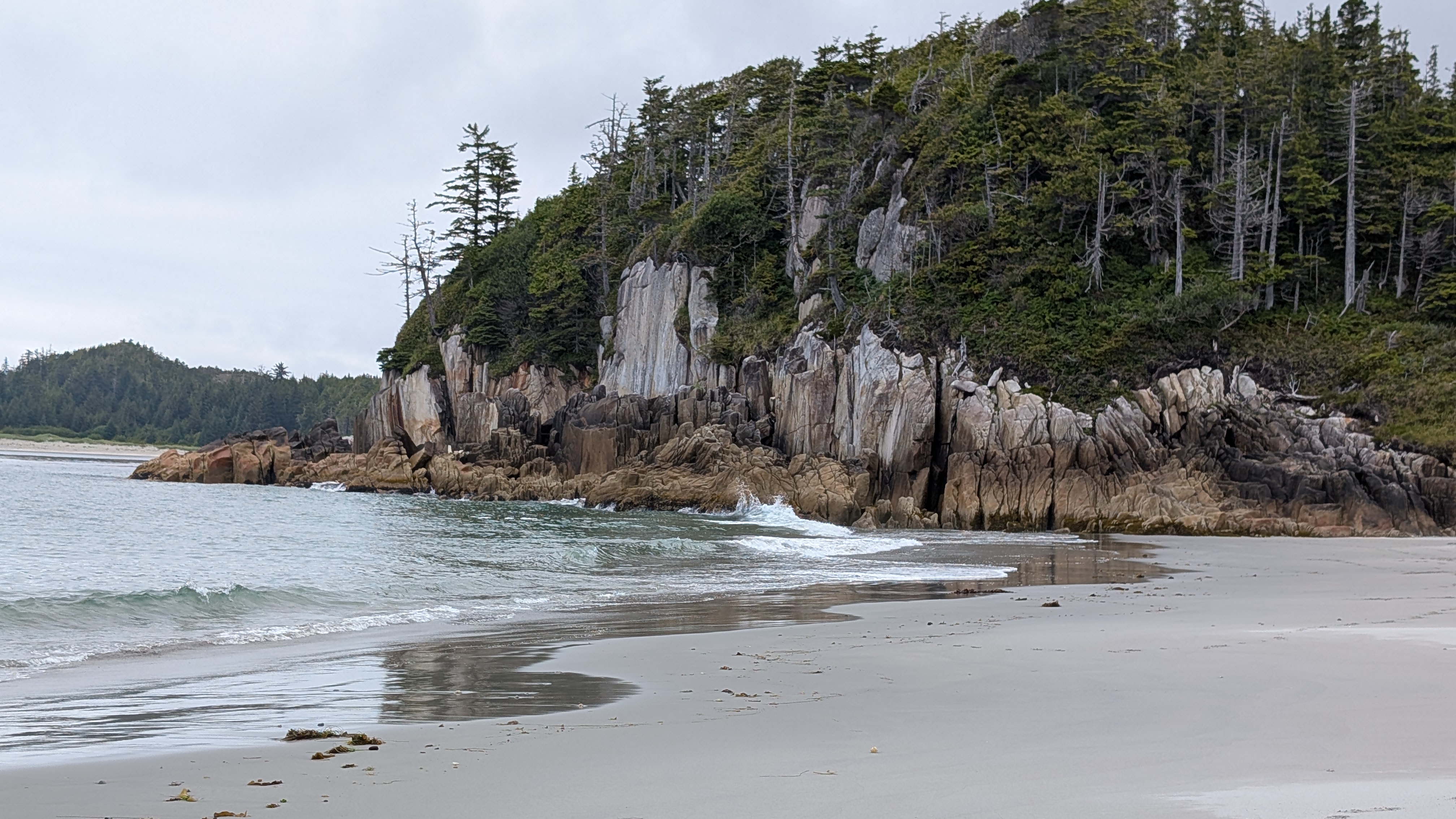 Dramatic rocky cliffs topped with evergreens meeting the sandy beach at Pruth Bay