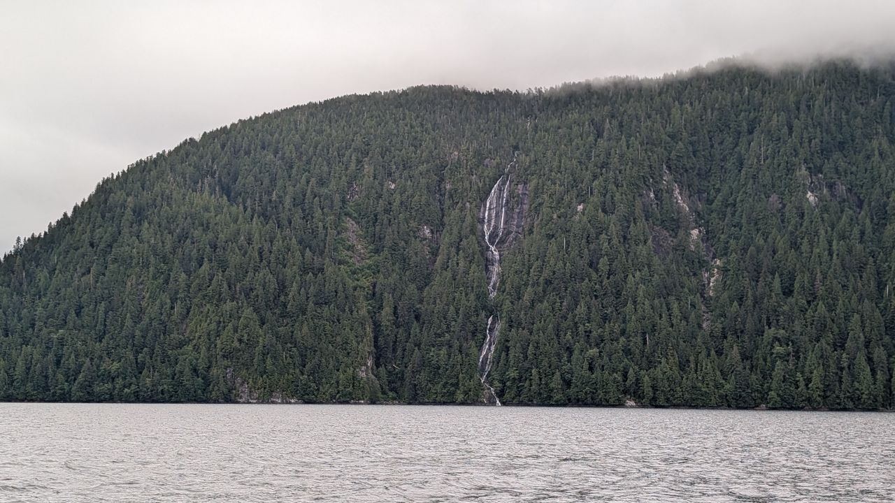 Waterfall cascading down a lush forested mountainside into the channel on an overcast day