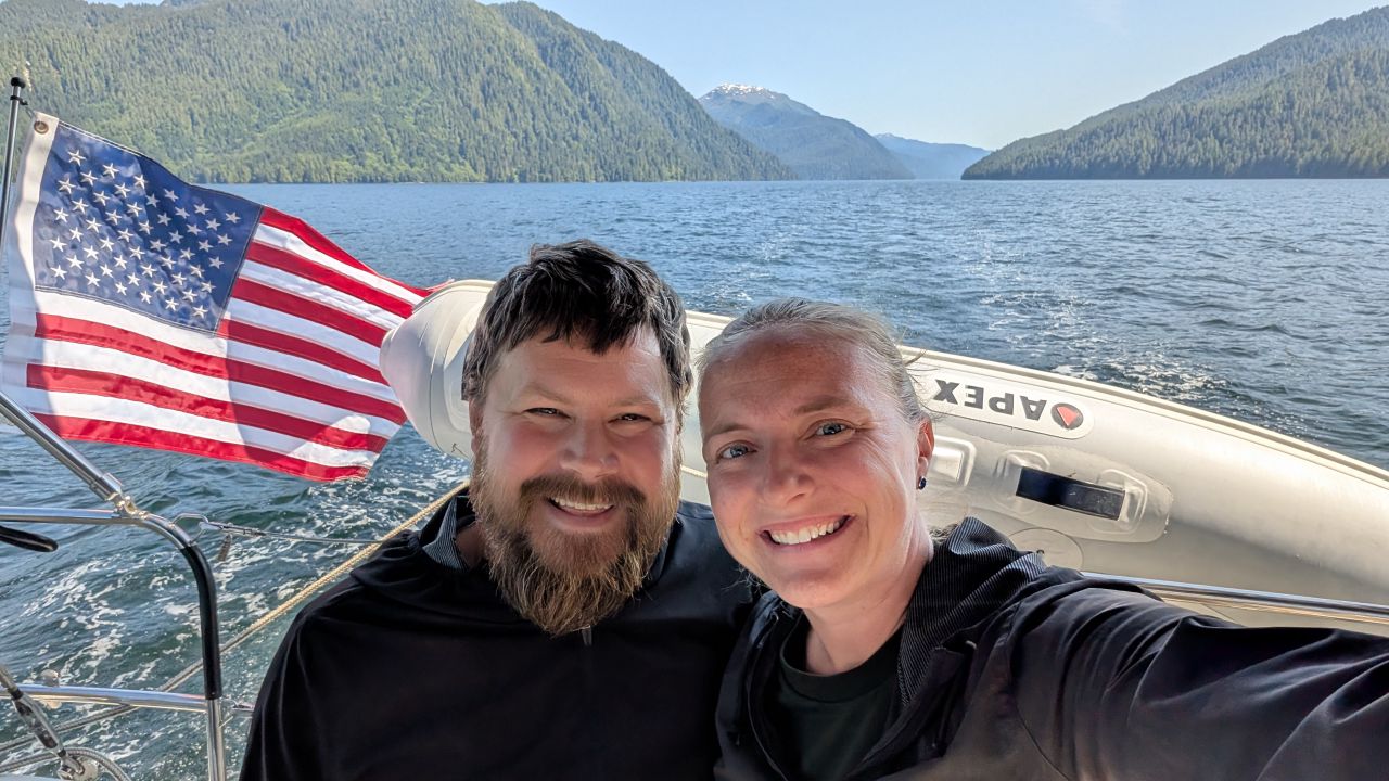 Rachel and Brian smiling for a selfie in the cockpit with mountains and American flag on the passage to Butedale
