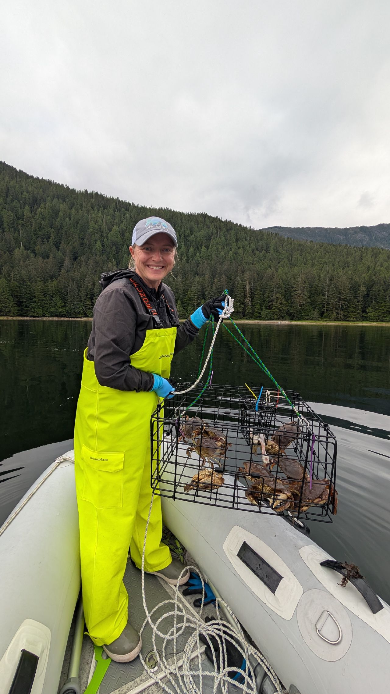 Rachel in yellow rain bibs proudly holding up the full crab trap with five crabs in the dinghy