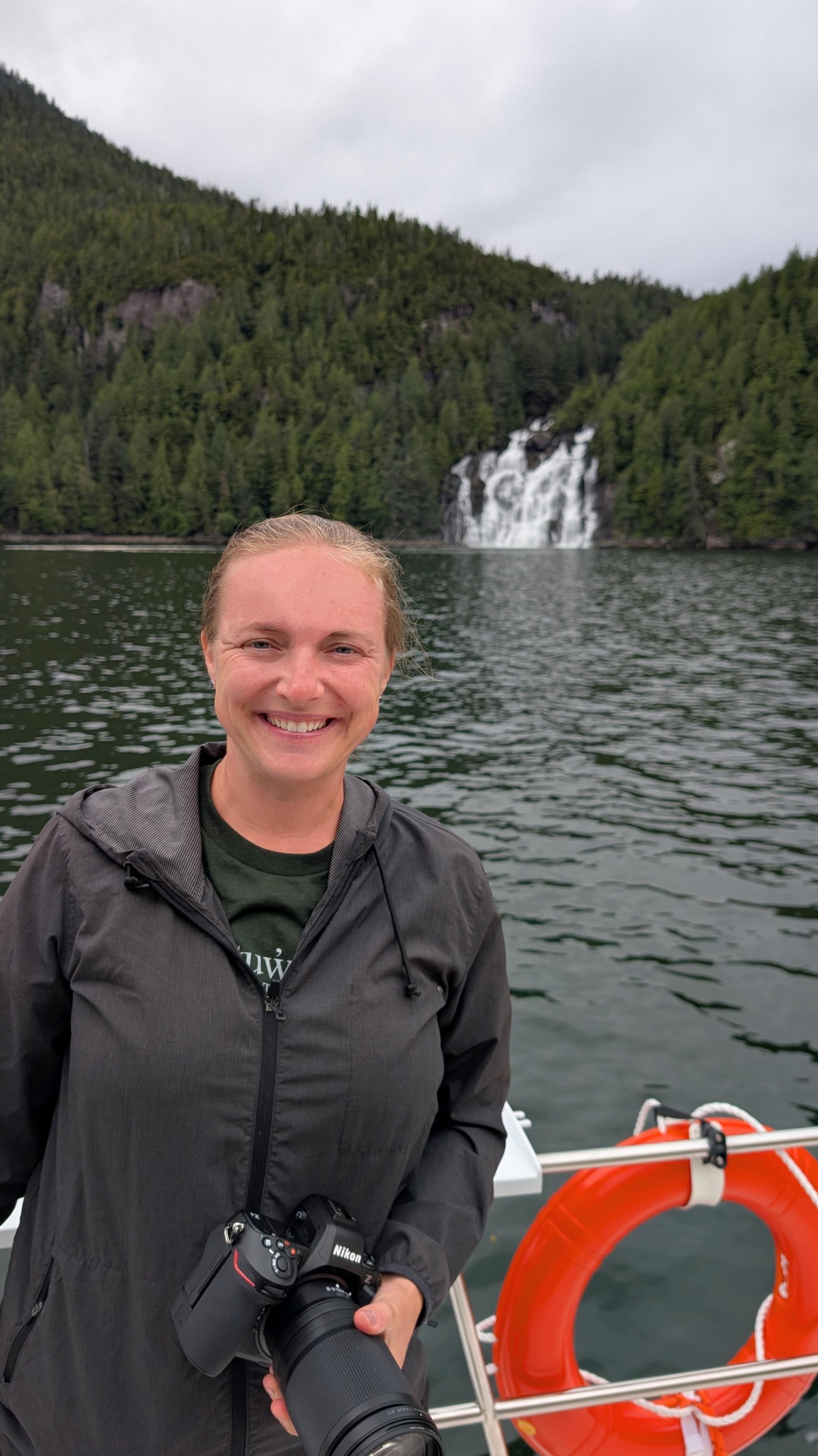 Rachel smiling with her camera and a life ring, the Poison Cove waterfall visible behind her