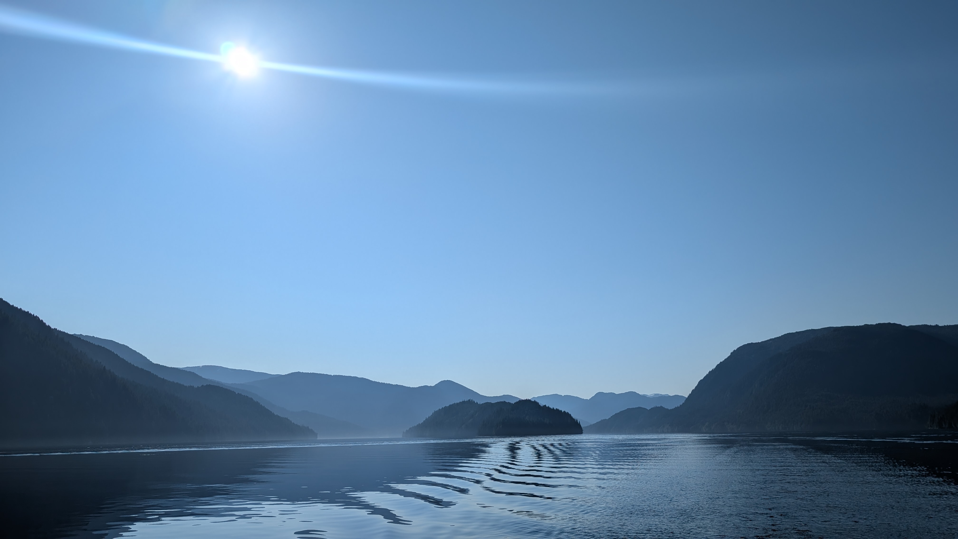 Hazy sun over layered mountain silhouettes and glassy water en route to Hartley Bay