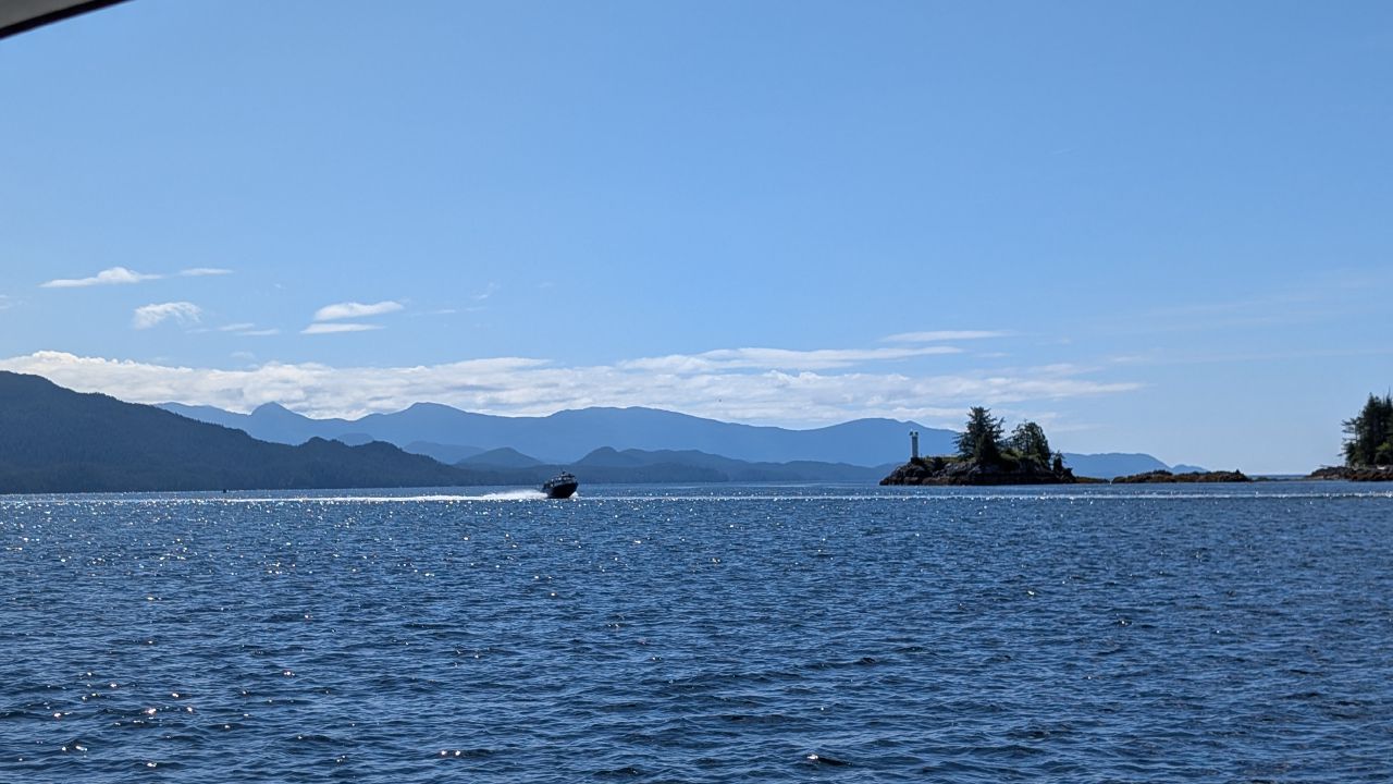 A boat motoring past a small island with a lighthouse on a sunny day with layered blue mountains in the background