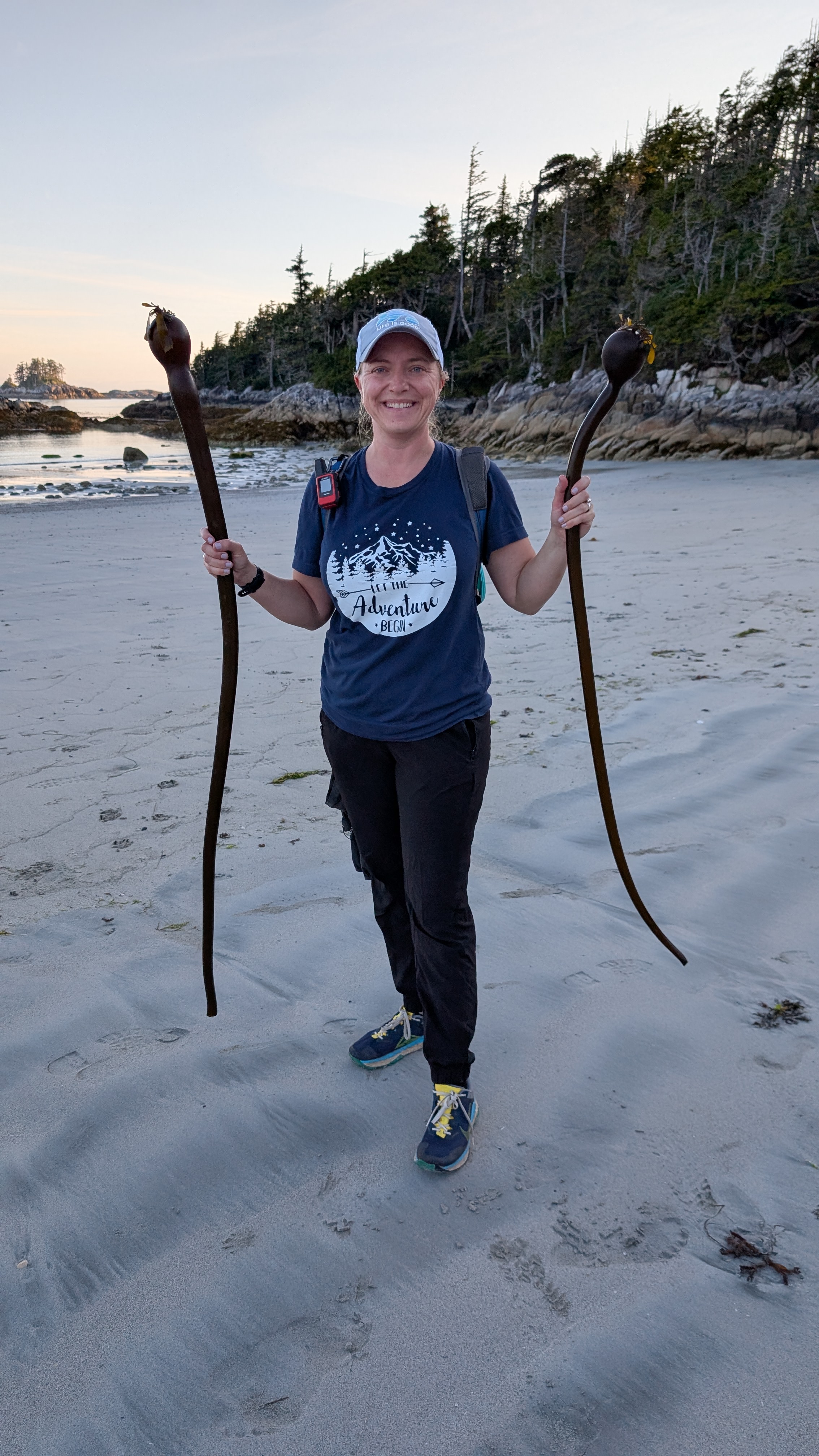 Rachel smiling and holding two large pieces of bull kelp on the beach at Pruth Bay