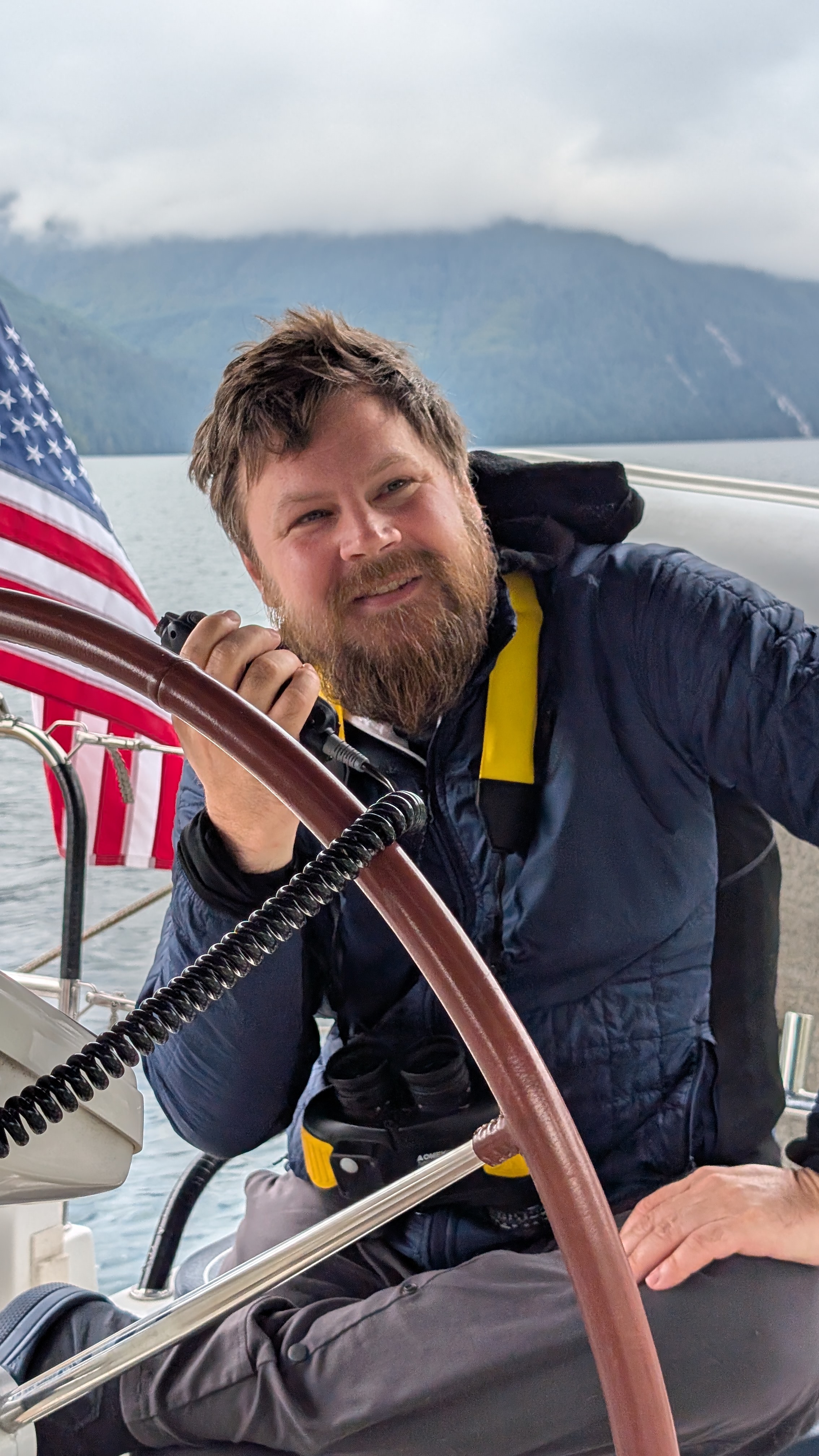 Brian smiling at the helm of SV Yahara with the American flag and cloud-covered mountains behind