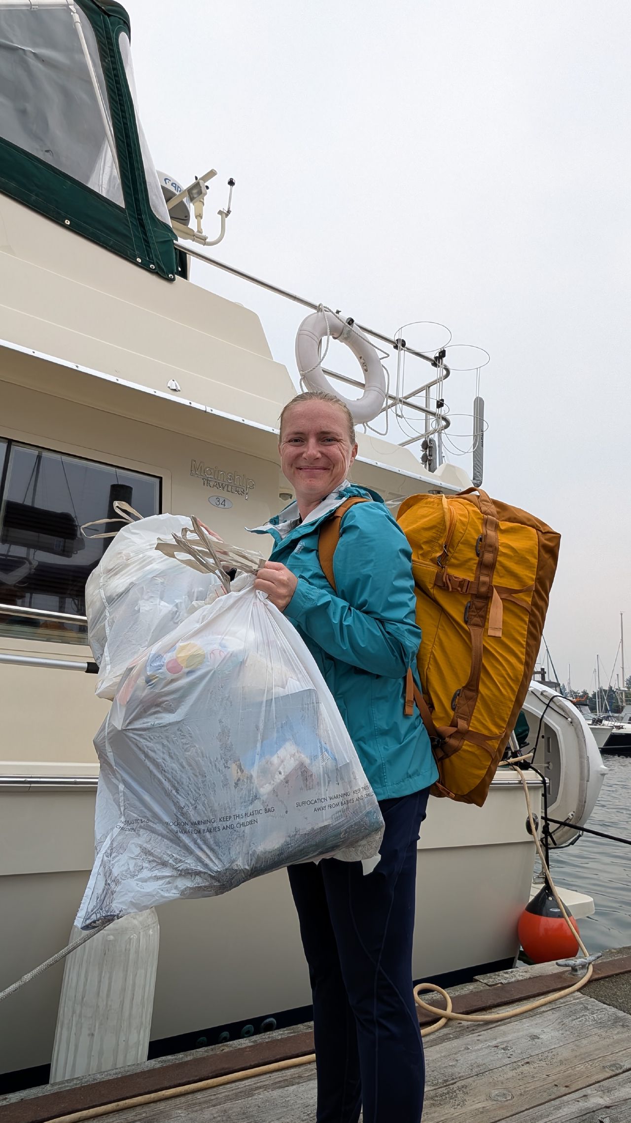 Rachel smiling on the dock carrying a large garbage bag and orange duffel after a month of remote cruising in Comox