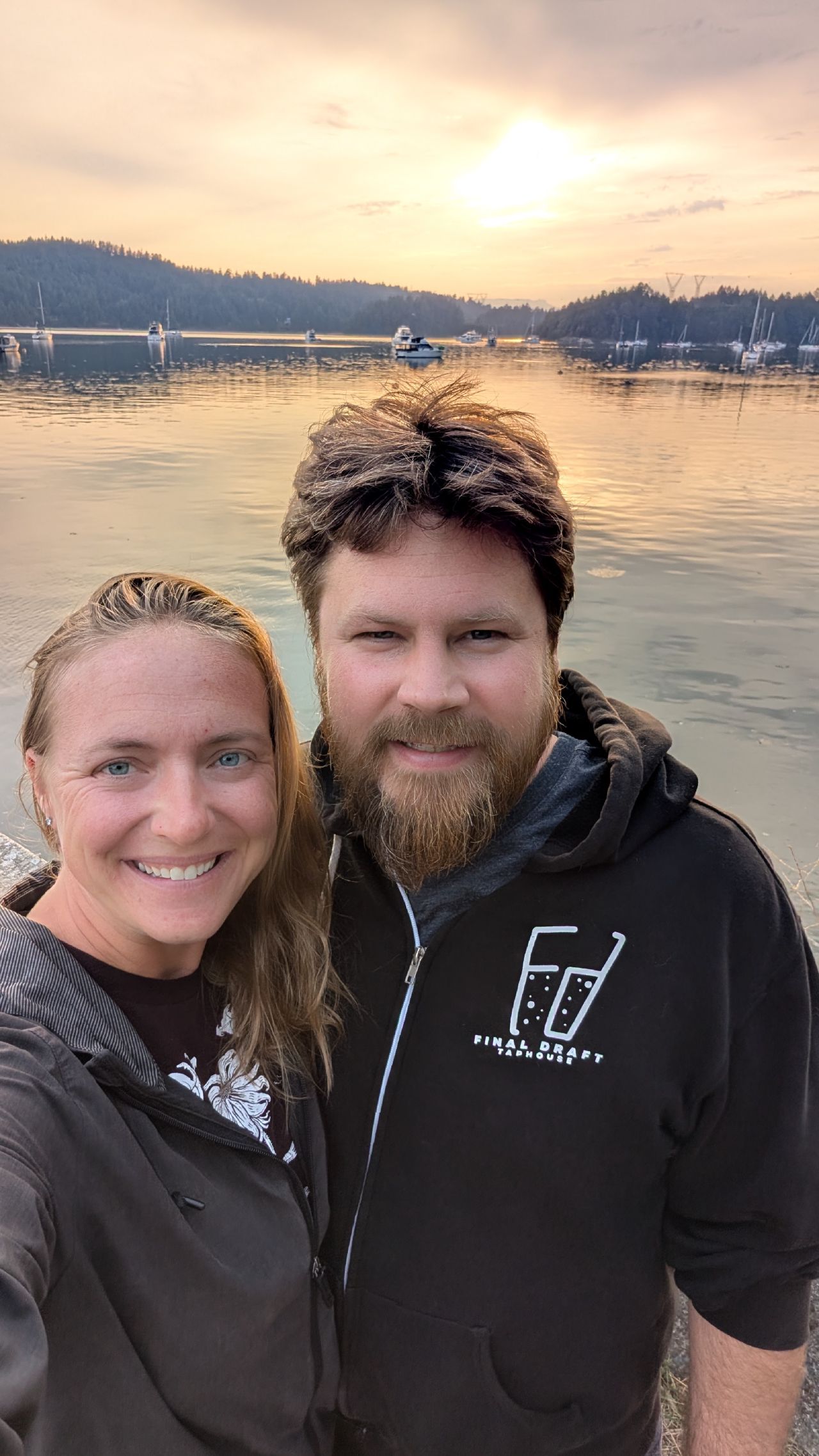 Rachel and Brian smiling for a selfie at sunset with boats anchored in Montague Harbour behind them