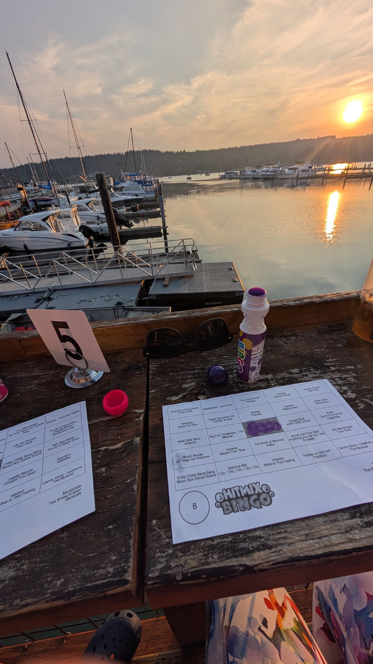 Disney music bingo cards and dabbers on a rustic bar table overlooking Poulsbo marina at sunset