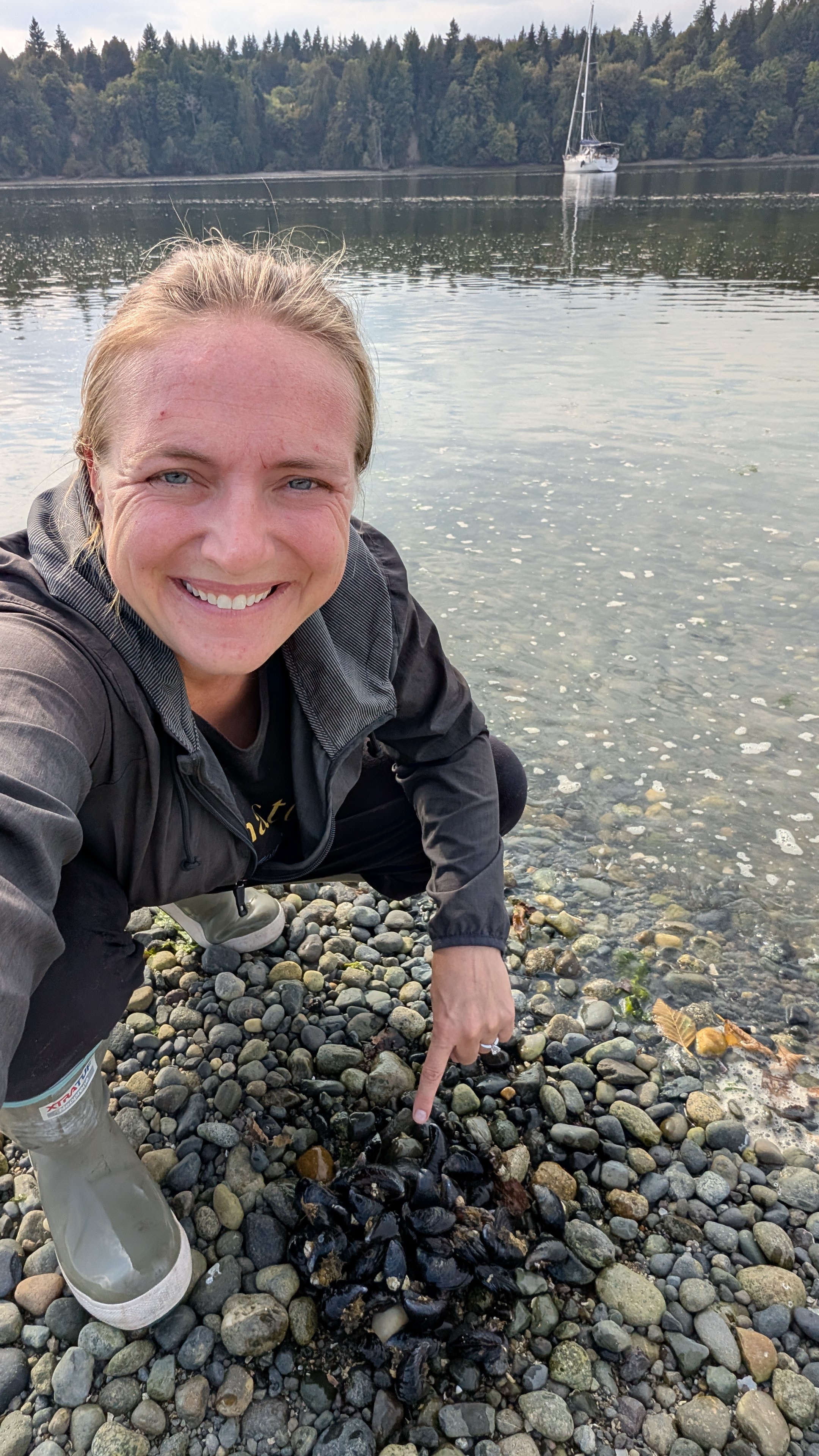 Rachel in rain boots pointing at mussels on the rocky beach with SV Yahara anchored behind her